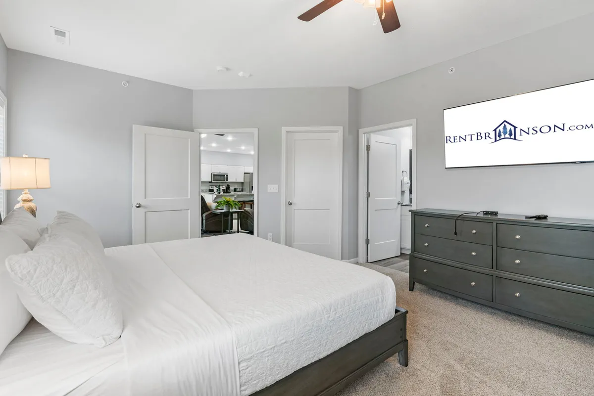 A calm and comfortable bedroom with a neatly made bed, dresser, and television. Doorways open to additional spaces, creating a sense of spaciousness while maintaining a restful, homey atmosphere.