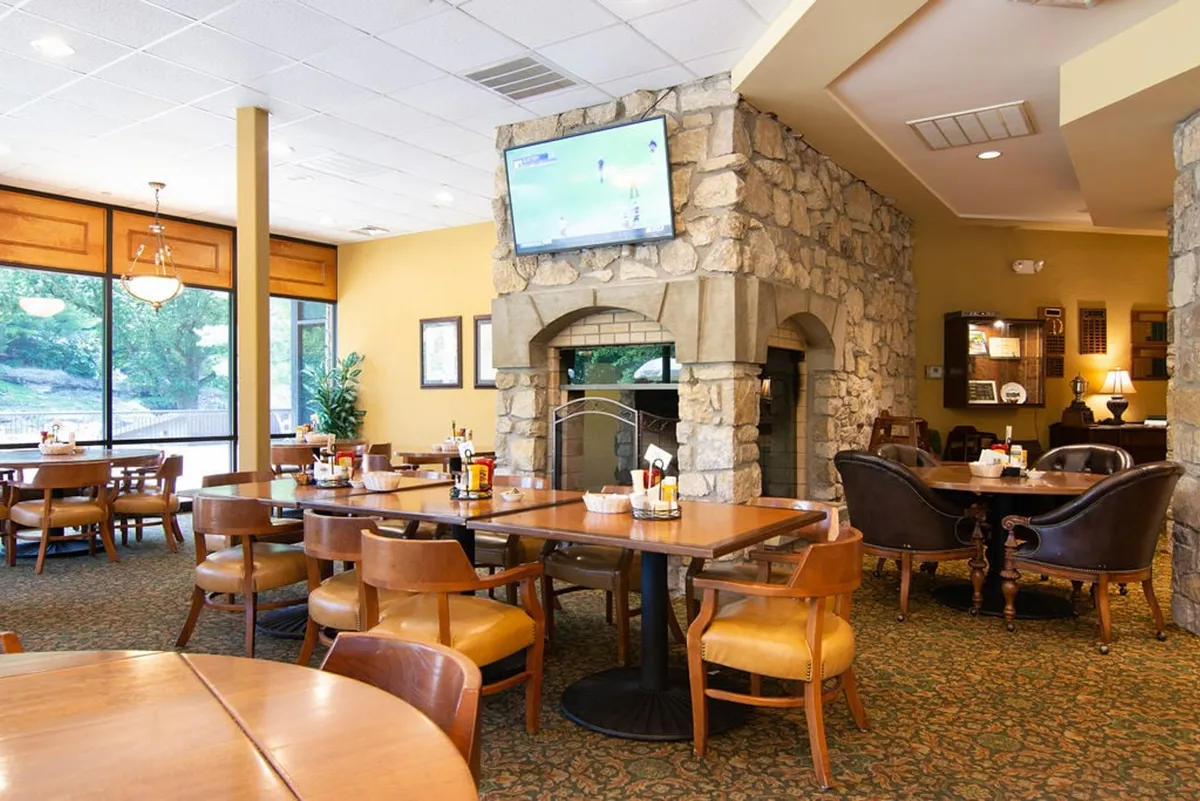 An inviting clubhouse interior with a large stone fireplace, rustic wooden tables, and expansive windows offering views outside. Comfortable seating and a television imply a place for post-game relaxation and socializing.