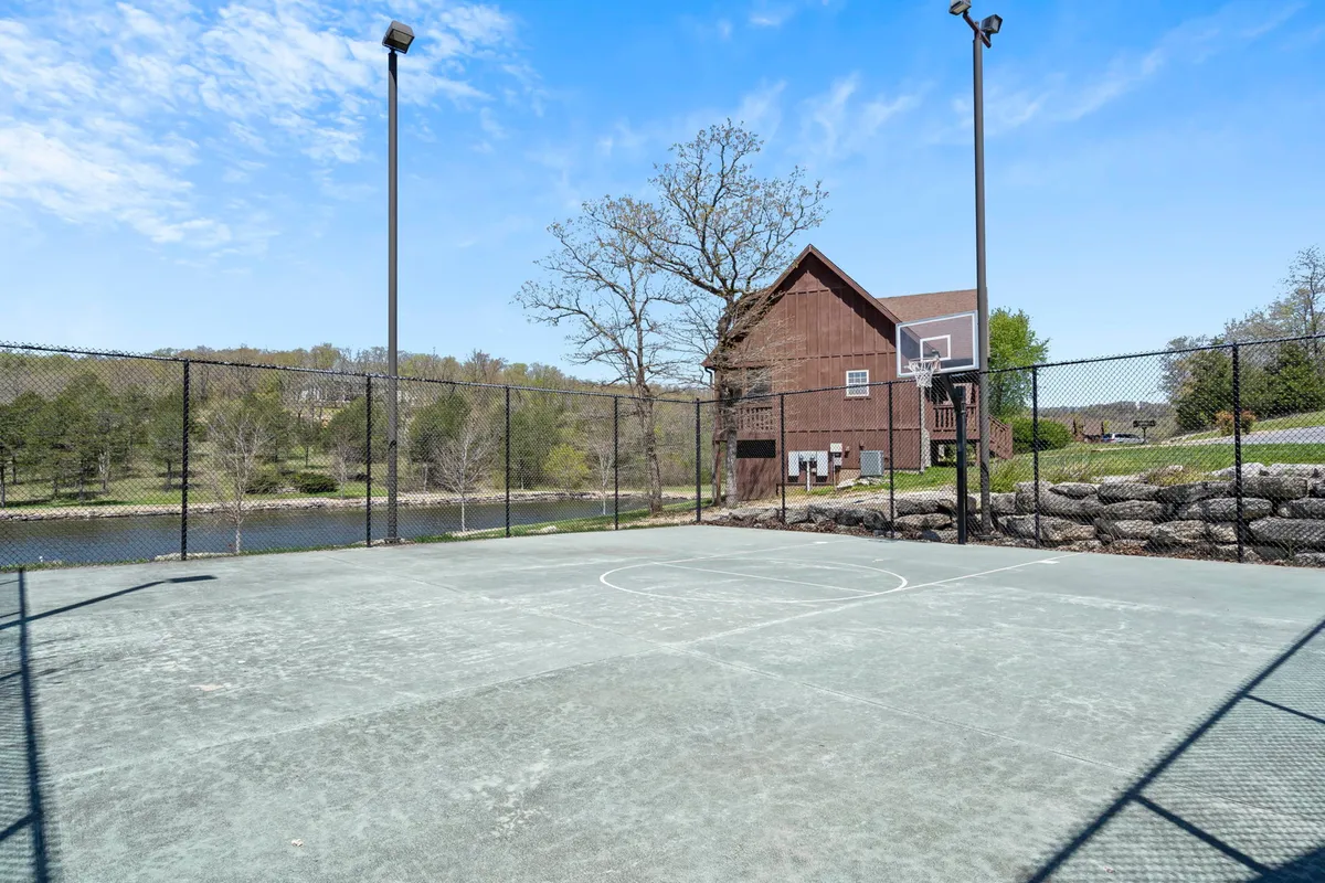 An outdoor basketball court framed by trees and overlooking a serene lake, with a house visible in the background. The scene balances active recreation with a natural, peaceful environment, inviting both play and relaxation.