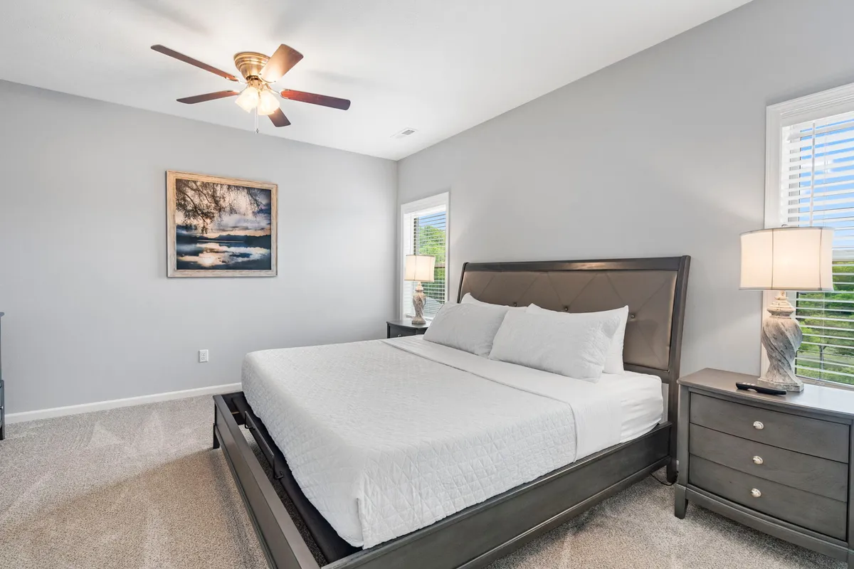 A spacious and inviting bedroom featuring a plush king-sized bed flanked by nightstands, soft artwork above the headboard, and large windows that bathe the room in natural light. The overall ambiance is tranquil and perfect for relaxation, suggesting a luxurious retreat after a day of activity.