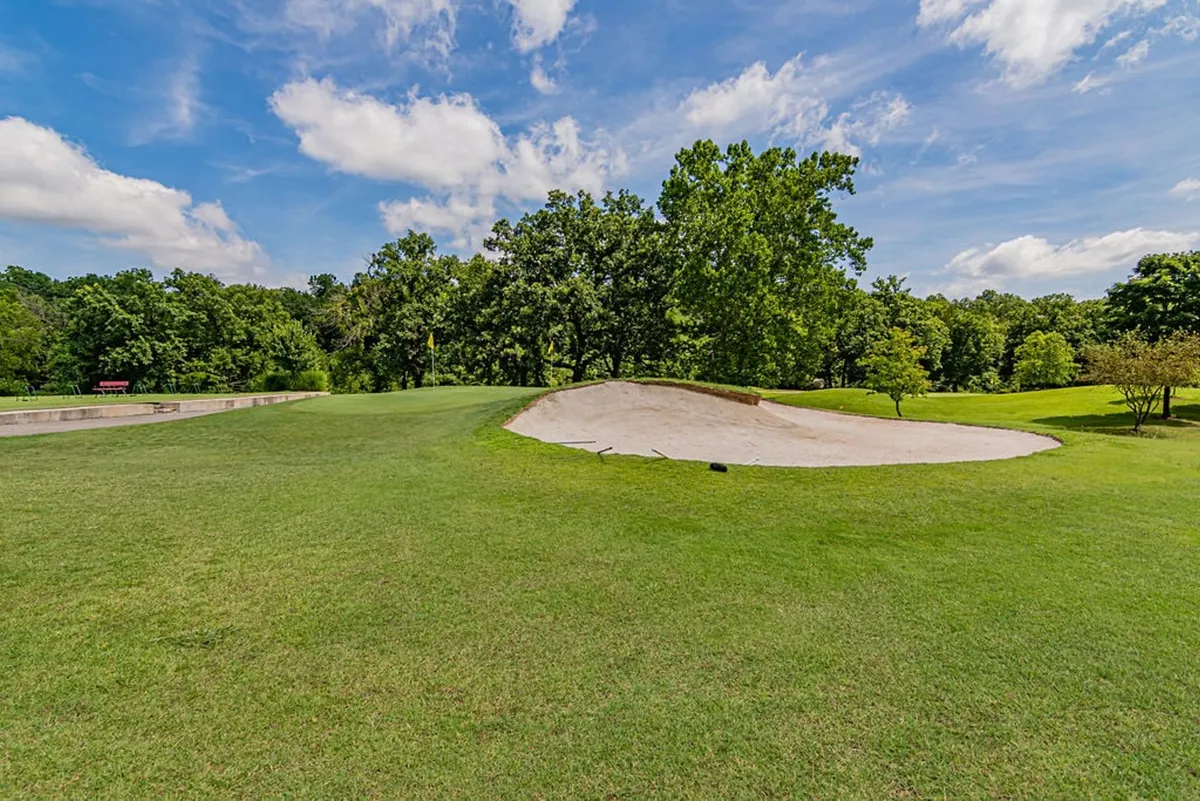 This image presents a wide view of a golf course, with a prominent sand trap, lush green fairways, distant trees, and a bright blue sky. The scene emphasizes the manicured beauty and tranquility of the golf grounds.