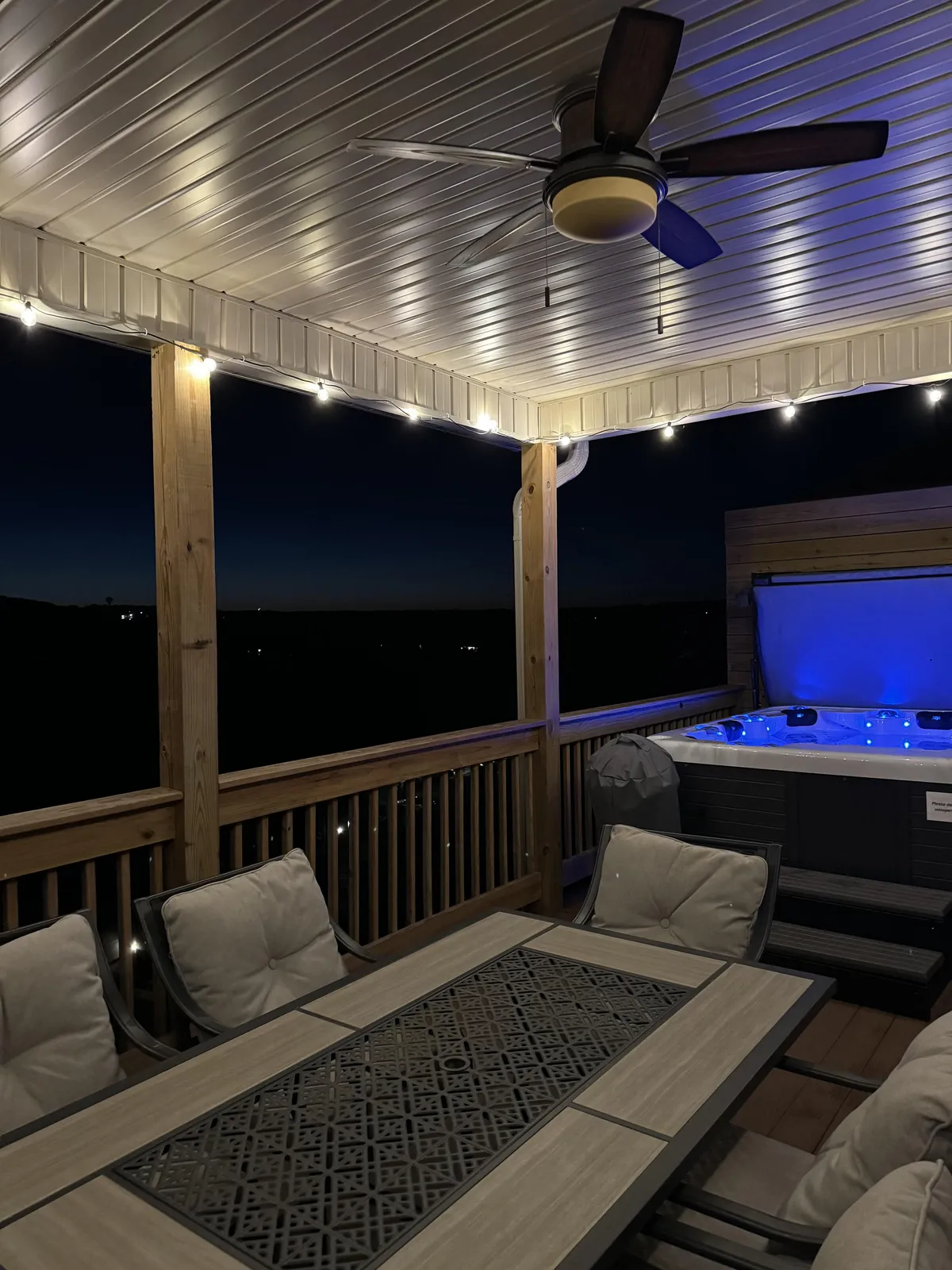 This image captures a private outdoor deck featuring a dining table, a prominent hot tub, ceiling fan, and string lights overhead. The hot tub is integrated into the space, inviting relaxation amid a tranquil outdoor atmosphere. Evening light suggests opportunities for stargazing or cozy gatherings.