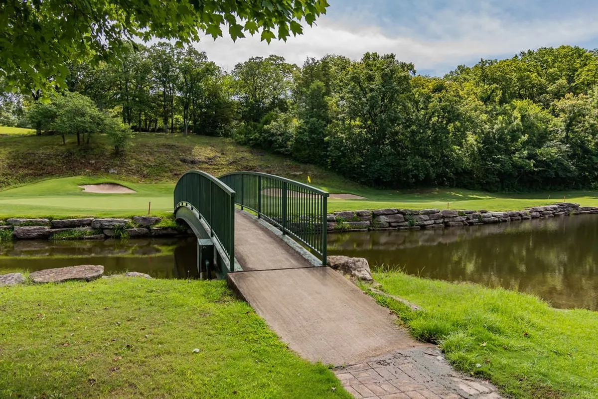 The image highlights a lush golf course scene with a green bridge stretching over calm water, surrounded by vibrant trees. The tranquil landscape is perfect for leisurely walks, golf outings, or simply enjoying peaceful moments in nature.