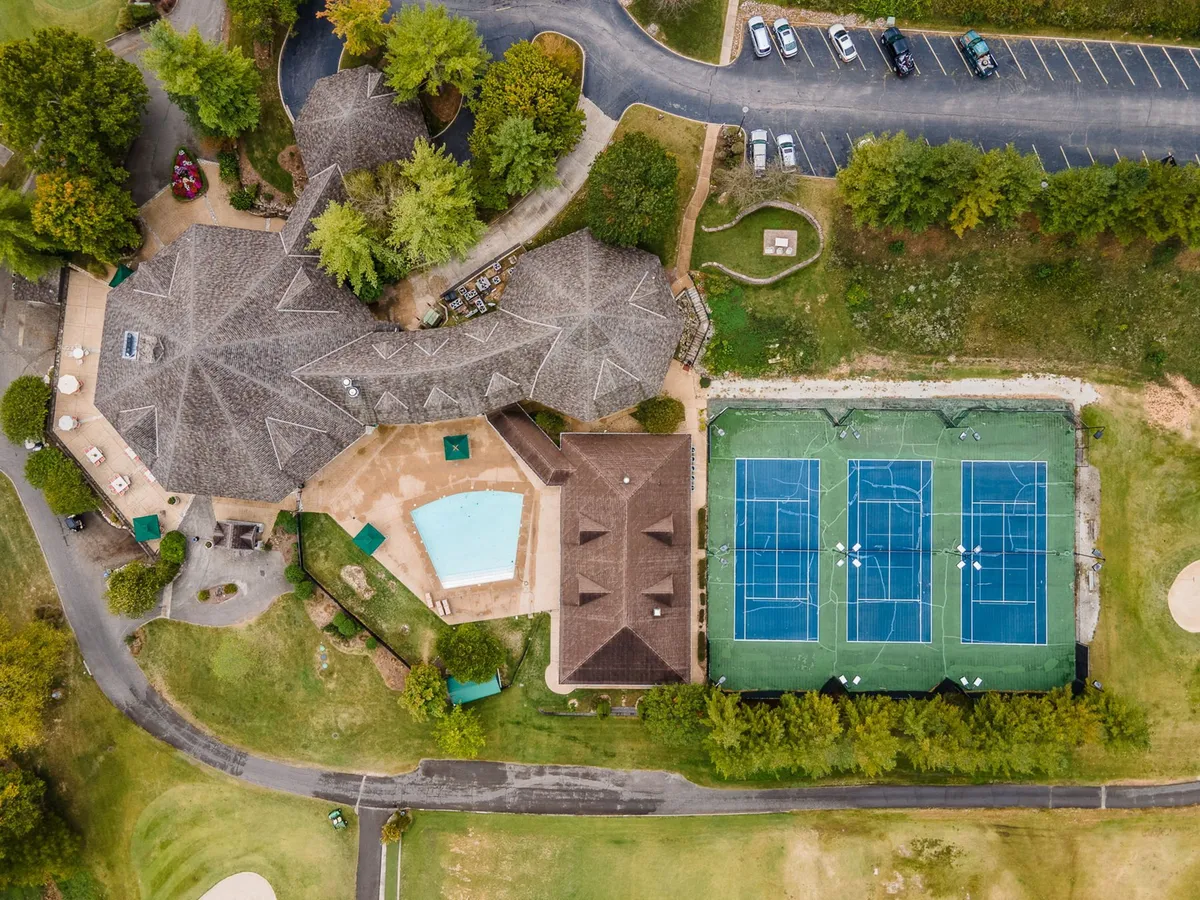 A lively exterior scene featuring a swimming pool surrounded by lounge chairs, adjacent tennis courts, a landscaped garden, and the clubhouse in the background. The area is bathed in sunlight, creating a vibrant and active setting that reflects community engagement and relaxation.
