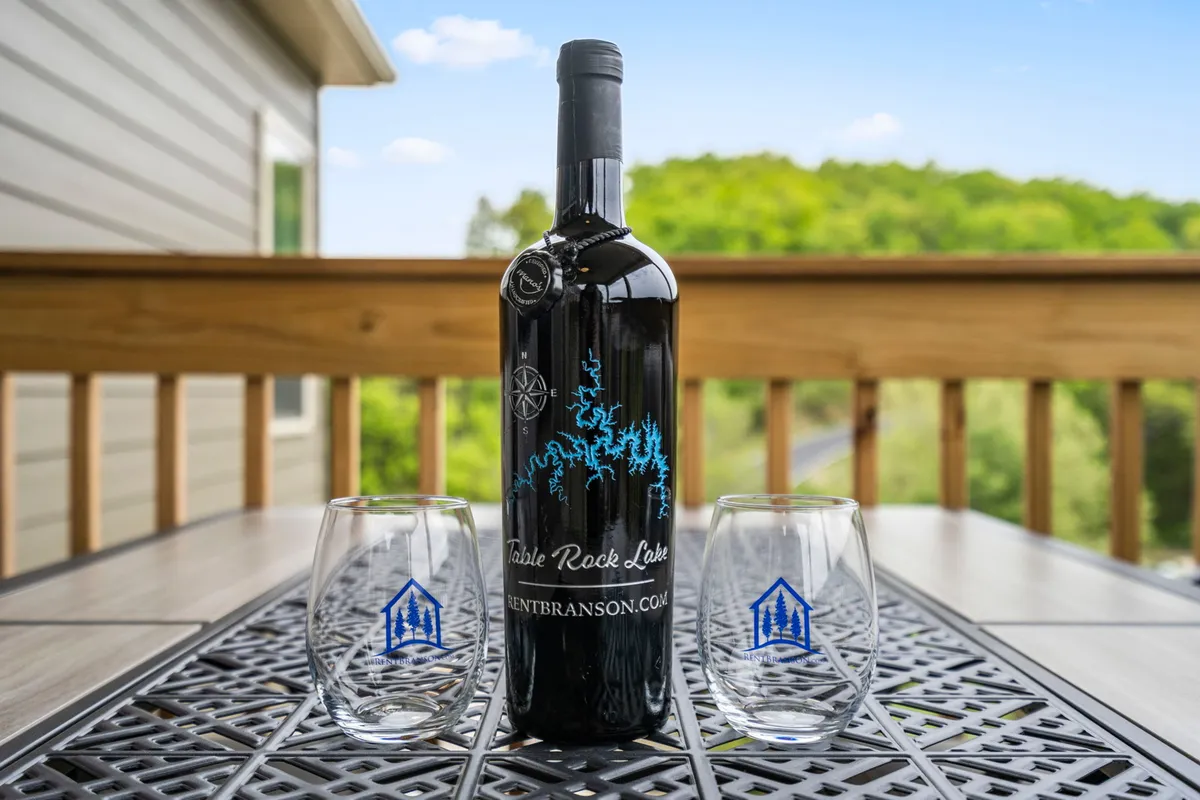 This image captures a serene outdoor setting with a wine bottle and two glasses placed on a wooden railing, overlooking lush green trees in the background. The scene evokes a sense of relaxation and intimacy, ideal for couples or friends unwinding together with drinks while enjoying nature.