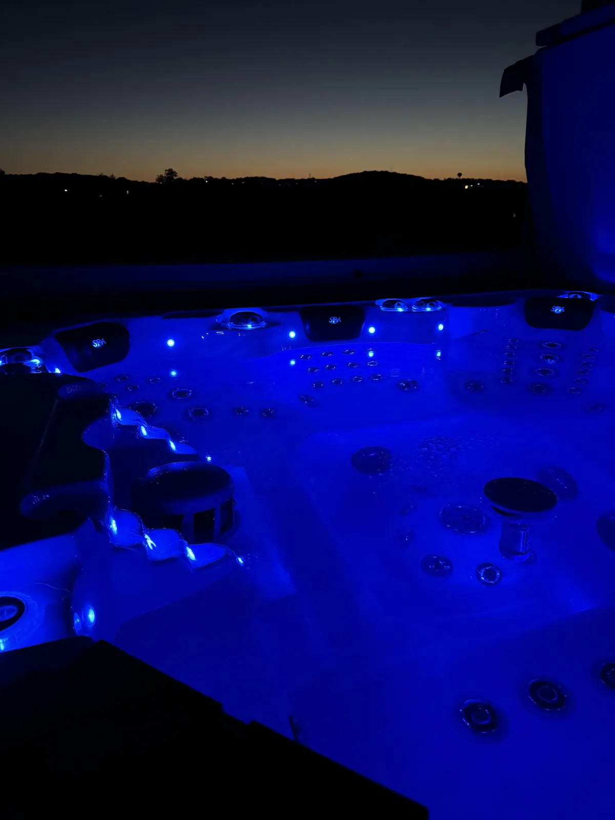 A close-up exterior shot of a hot tub illuminated by striking blue LED lights, emphasizing the bubbling water surface. The night setting and calming lights enhance the feeling of immersive relaxation and intimacy.